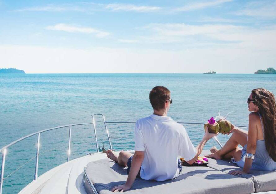 Two people sit on a yacht overlooking a blue, open ocean on a sunny day with several islands in the distance.