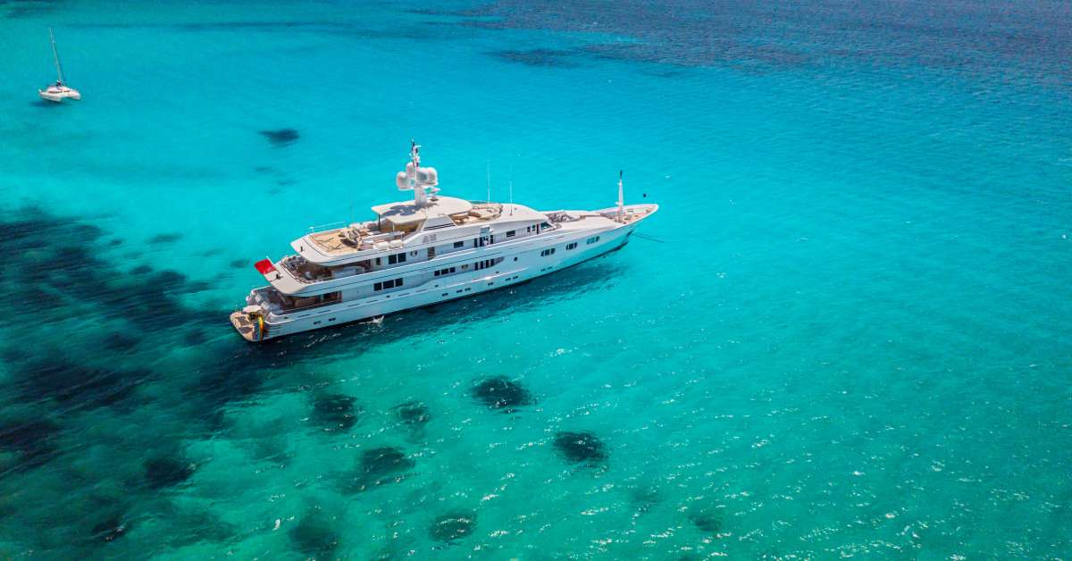 A massive yacht with multiple decks is sitting still in the bright blue waters of the ocean and the sun is overhead.