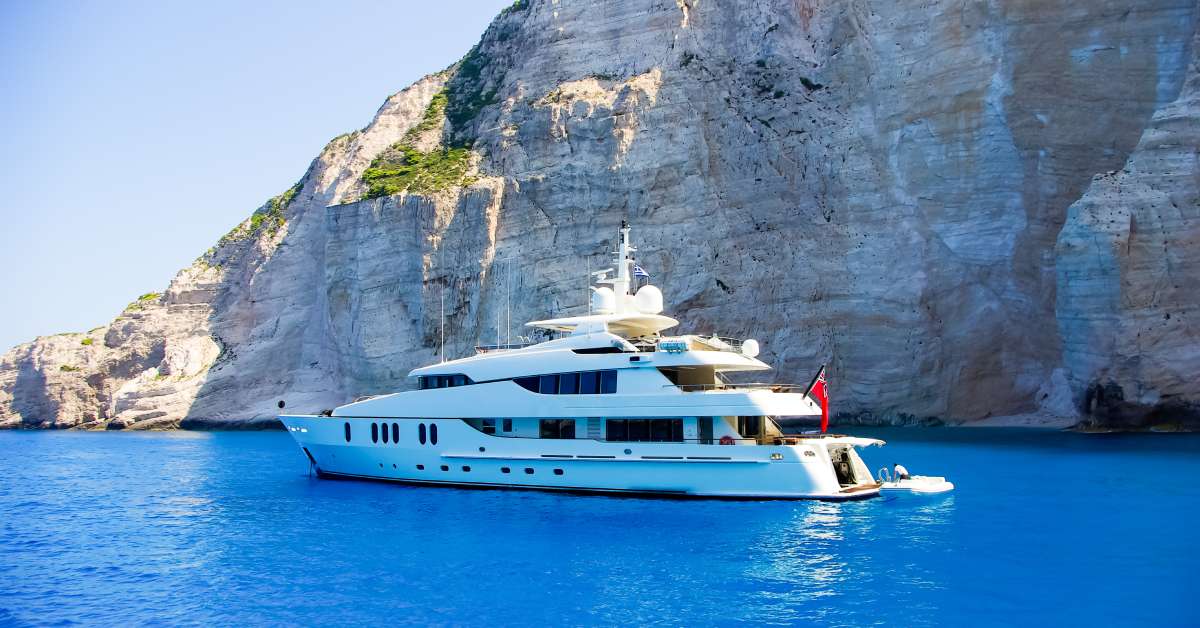 A yacht sits on vibrant blue waters in front of a large rock face. A crew member boards a small boat at the back of the yacht.