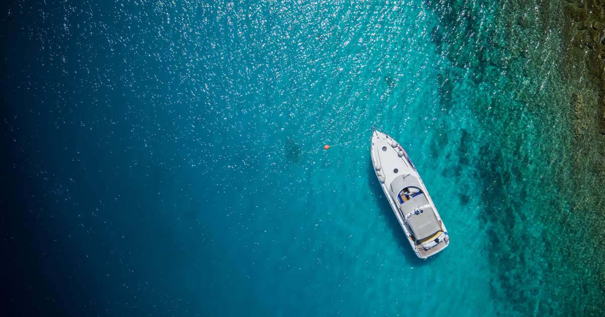 A white yacht sits still on the ocean. The blue water is so clear that you can see the seabed under the boat.