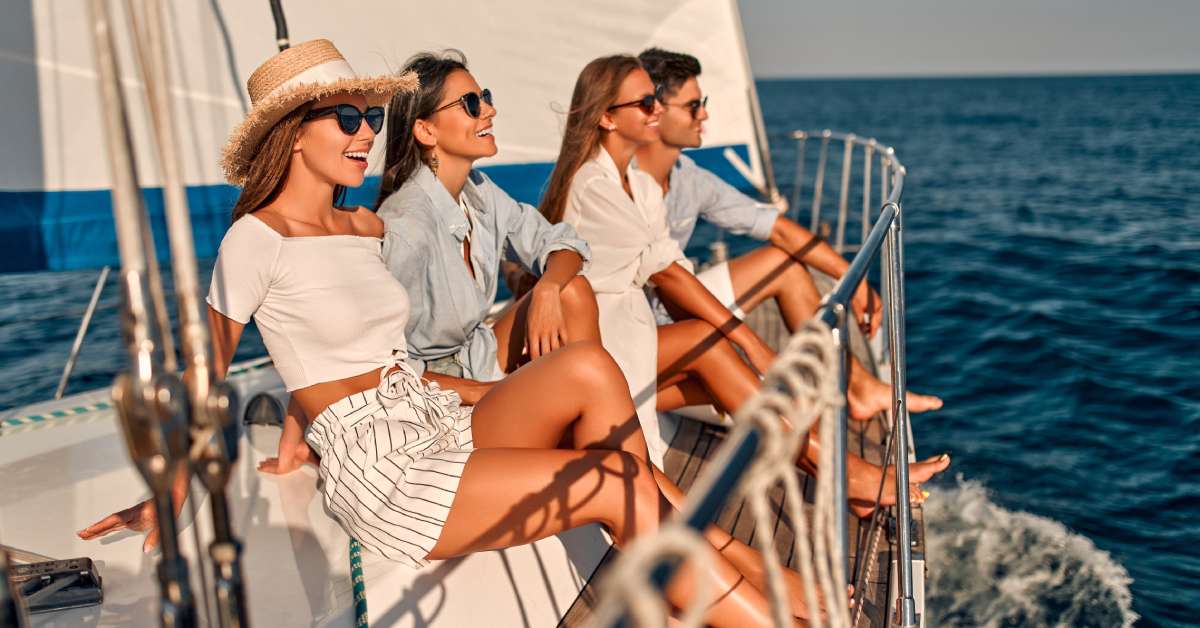 Four friends wearing sunglasses, smiling and sitting near the railing of their yacht to watch the ocean.