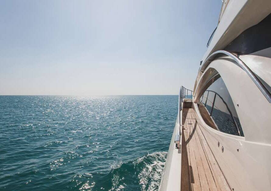 The side of a large yacht with a wooden deck and a metal railing. It sails smoothly on the ocean on a sunny day.