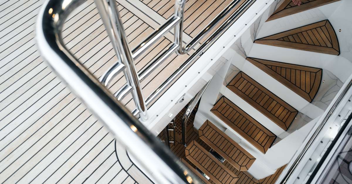 A wood floor on the second level of a yacht leads to stairs that go to a lower level. The stairs have a metal railing.