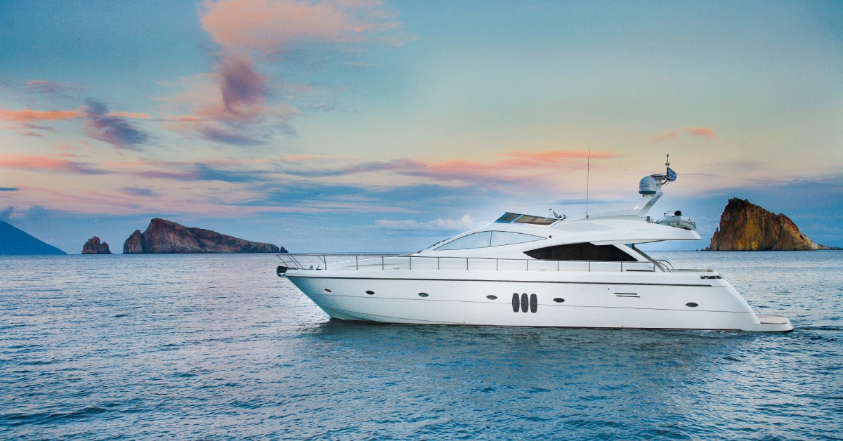 A yacht is sitting in the ocean, and two large pieces of land are visible in the distance. The sky is blue and orange.