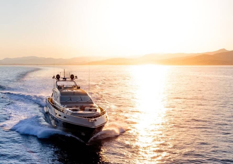 A yacht is moving quickly in the ocean, leaving big waves in its wake. It is moving away from the shore and the sun.