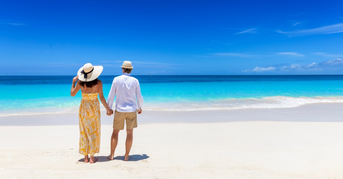 A couple wearing hats and standing on the beach with their backs to the camera. They watch the bright blue ocean's waves.