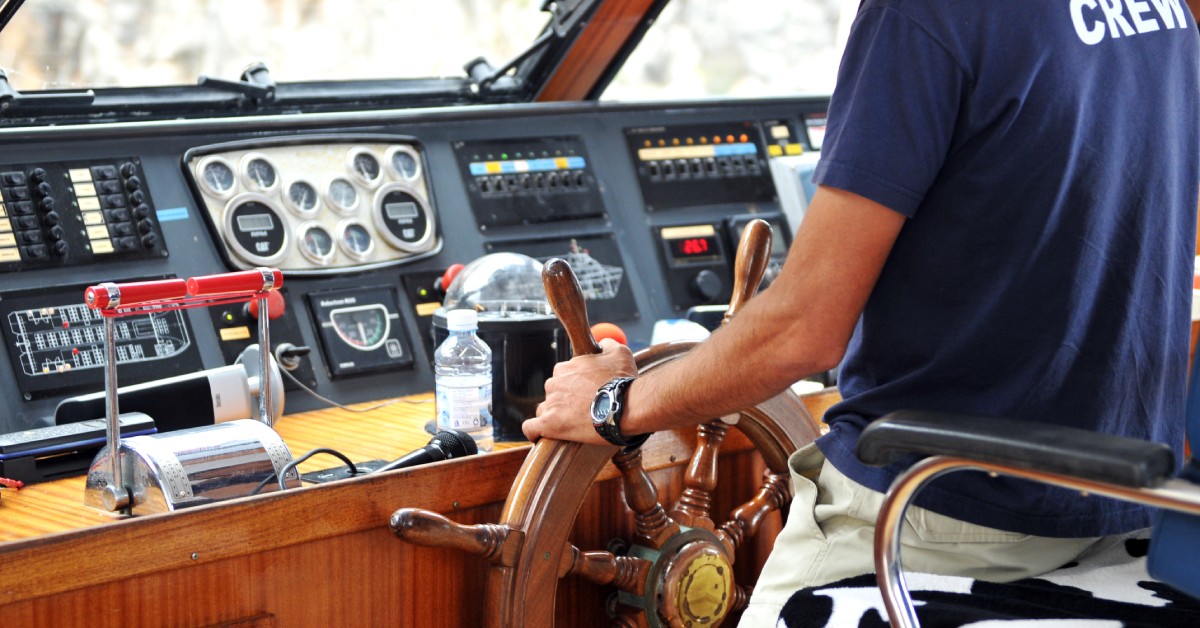 A person wearing a watch and a dark blue shirt that reads “Crew” on the back is operating a boat's steering wheel.