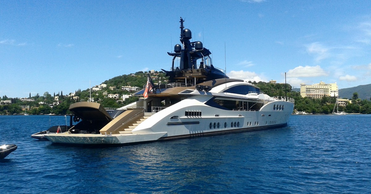 A massive white yacht sailing toward the nearest land, which is full of green trees and white buildings.