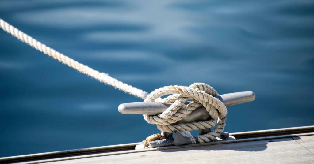 The rope for a boat is tied into a knot on a metal anchor point at the edge of a pier during the day.