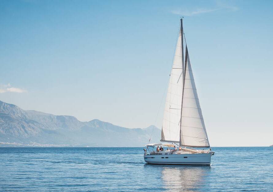 A white yacht with a massive sail sitting in the middle of the ocean. A large mountain is visible in the distance.