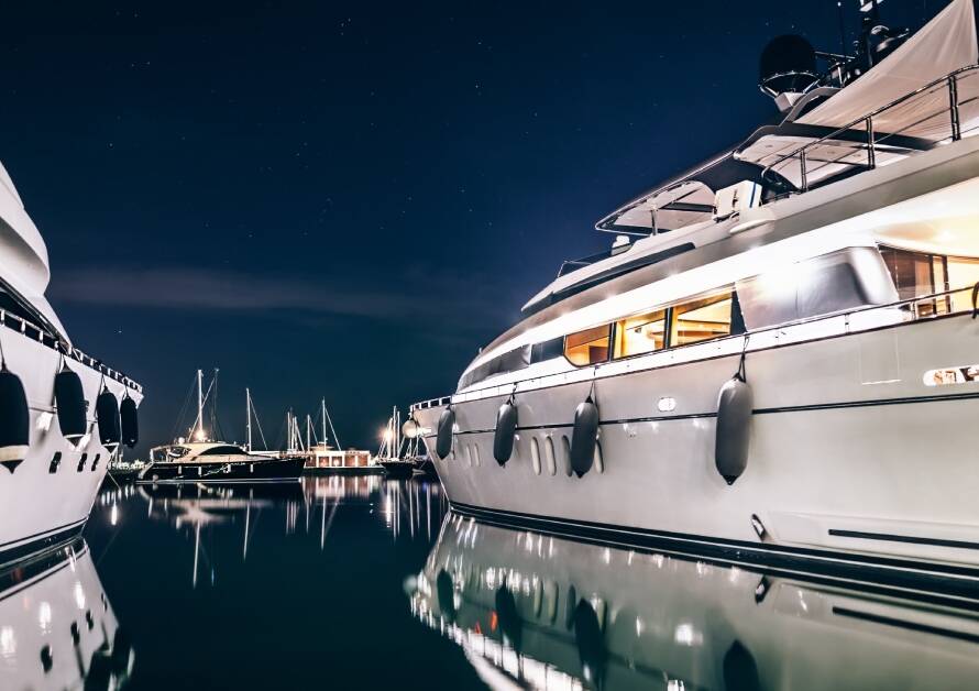 Two yachts sitting beside each other at night. Several other boats are visible in the distance behind the yachts.