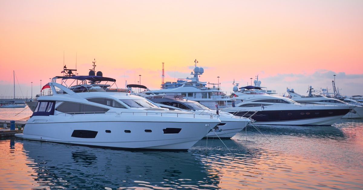 Six massive yachts floating in the water side-by-side while the sun sets, turning the sky bright orange.